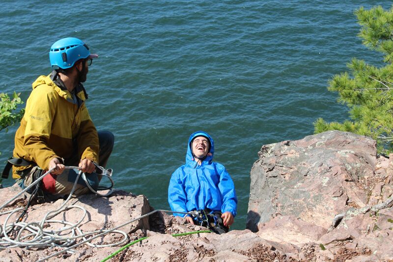 The image shows two people on a rocky cliff overlooking a body of water. One person is wearing a yellow and brown jacket and a blue helmet, holding climbing ropes. The other person is wearing a blue hooded jacket and is sitting on the rocks. The background features a large body of water and some greenery.